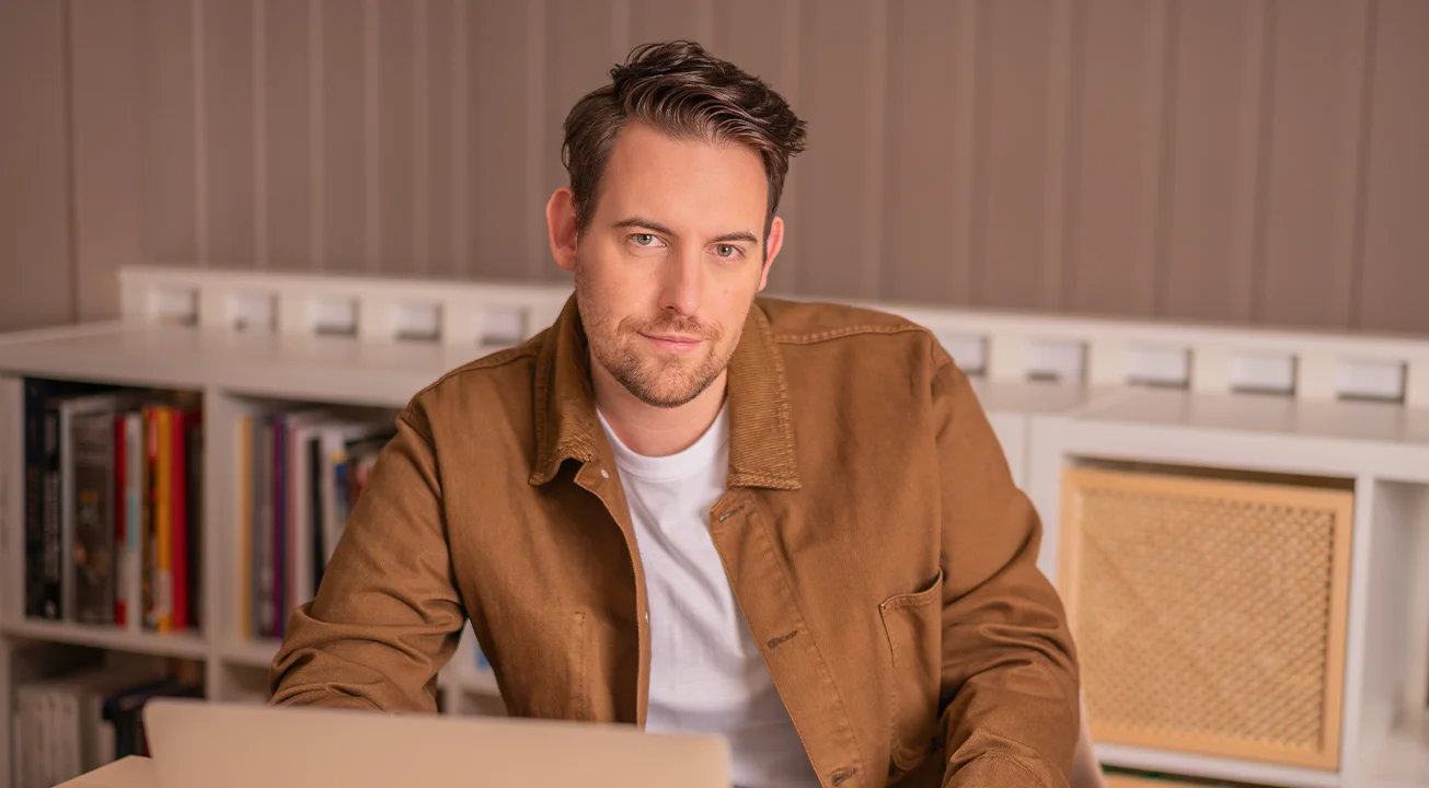 Bence Székely, the Strategy Lead of Frem Digital sits at a desk with a laptop, looking at the camera. Shelves with books and decor are in the background.