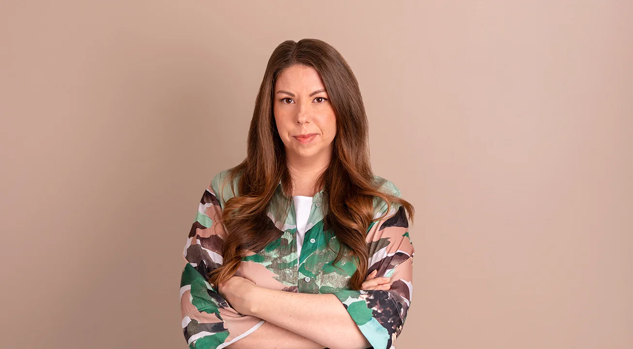 Orsi Imreh, co-founder of Frem Digital, stands in front of a beige background. She looks at the camera with her arms crossed, wearing a green patterned top.